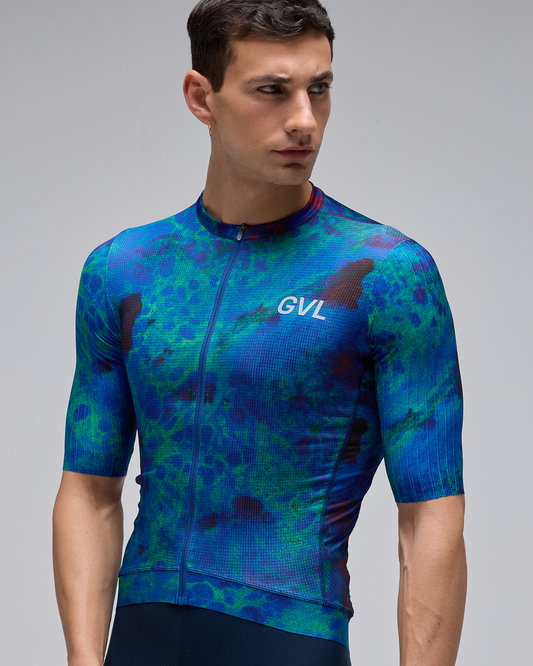 A man wearing a short-sleeve full-zip cycling jersey with a blue and green tie-dye pattern and a white GVL logo on the chest, set against a solid gray background.