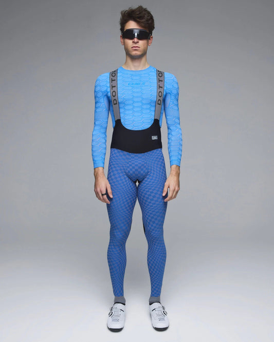 A model wearing a full-body blue patterned cycling suit with gray shoulder straps and a black waist panel, standing against a neutral gray studio background.