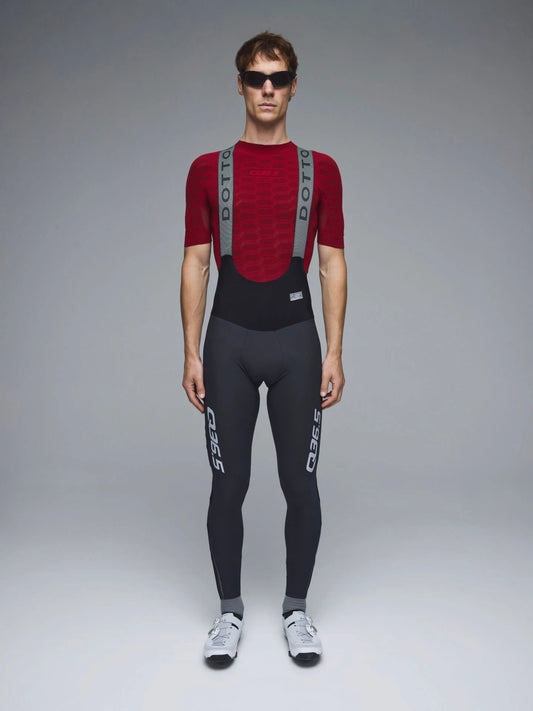 A male model wearing a red and black Dotti cycling bib short-sleeve jersey with gray shoulder straps and black bib shorts with white branding, standing against a neutral gray studio background.