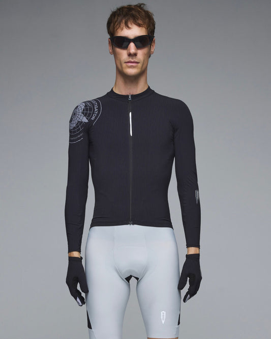 A male model wearing a black long-sleeve cycling jersey with a subtle graphic on the right shoulder, paired with light gray cycling shorts, black gloves, and sunglasses, against a solid gray studio background.