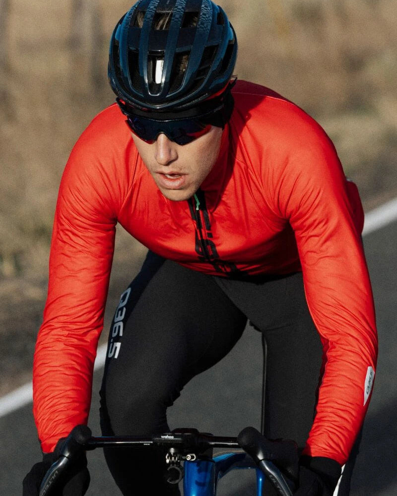 A cyclist in a bright red long-sleeve jersey and black cycling tights leans forward on a road bike outdoors. The rider is wearing a black helmet and dark sunglasses.