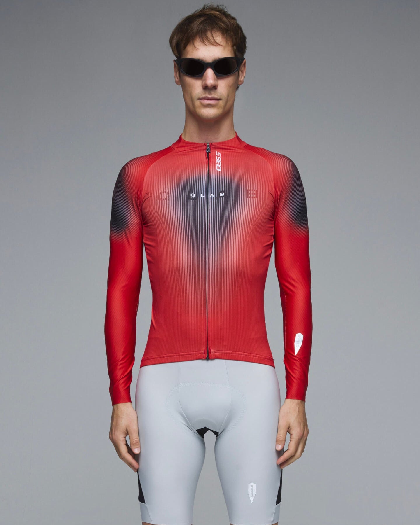 A male model wearing a red long-sleeve cycling jersey with a black-to-gray gradient chest design and light gray cycling shorts, standing against a neutral gray studio background.