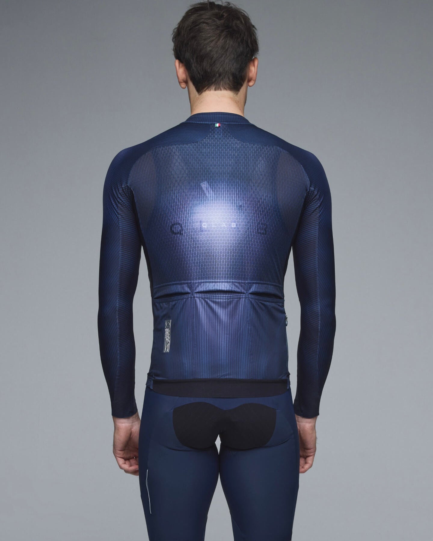 Rear view of a man wearing a dark navy blue long-sleeve cycling jersey and matching shorts with a black rear pad, against a solid gray studio background.