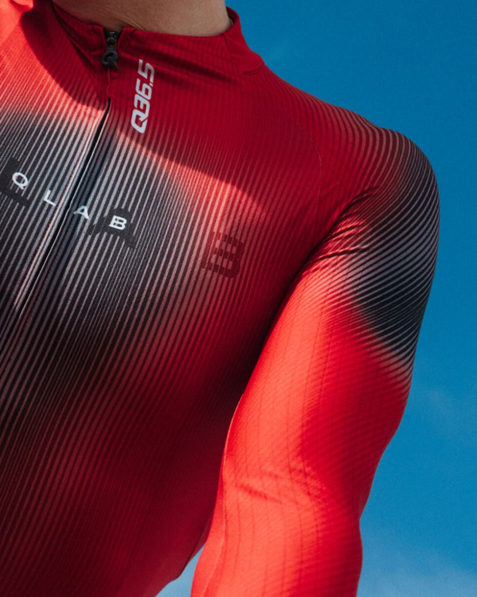 A close-up of a cyclist wearing a red and black ribbed aerodynamic jersey with a front zipper, holding a handlebar against a bright blue sky. The brand name 'G-1000' is visible on the chest.