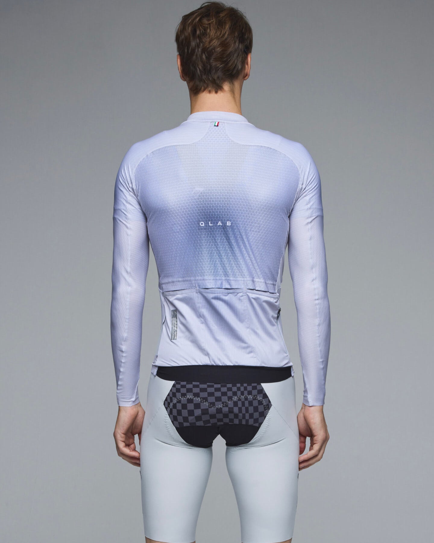 Rear view of a male model wearing a light blue-gray long-sleeve cycling jersey with a subtle gradient and textured pattern, paired with light gray shorts and dark patterned cycling briefs, against a solid gray studio background.
