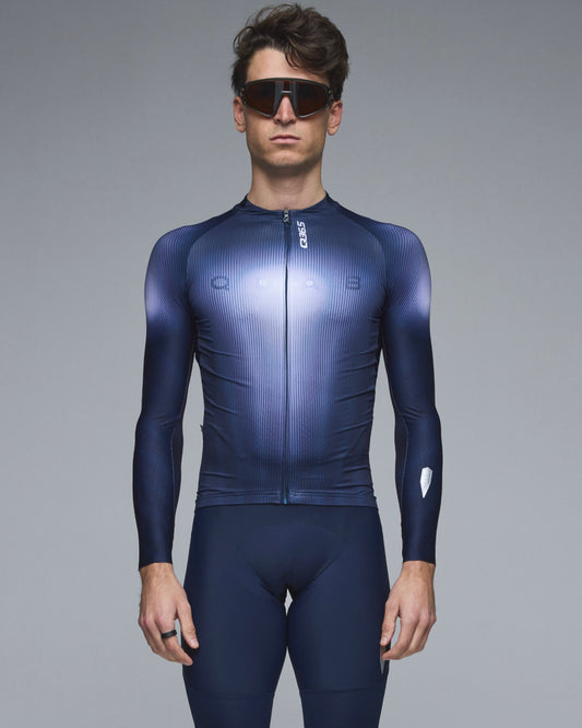 A male model wearing a dark blue and navy long-sleeve cycling jersey with matching shorts, black sunglasses, and a ring, standing against a solid gray studio background.