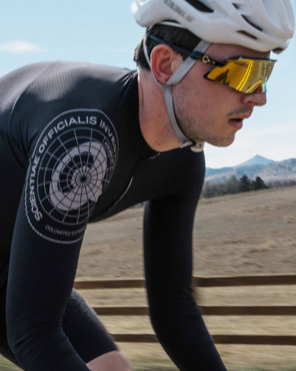 A cyclist in a black full-body cycling suit with a white circular logo on the shoulder, wearing a white helmet and yellow mirrored sunglasses, riding outdoors on a road with a fence and hills in the background.