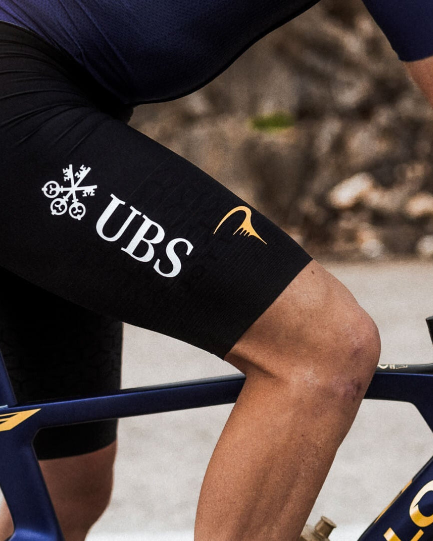 Close-up of a cyclist wearing black UBS bib shorts with gold and white logos, riding a dark blue and gold road bike outdoors.