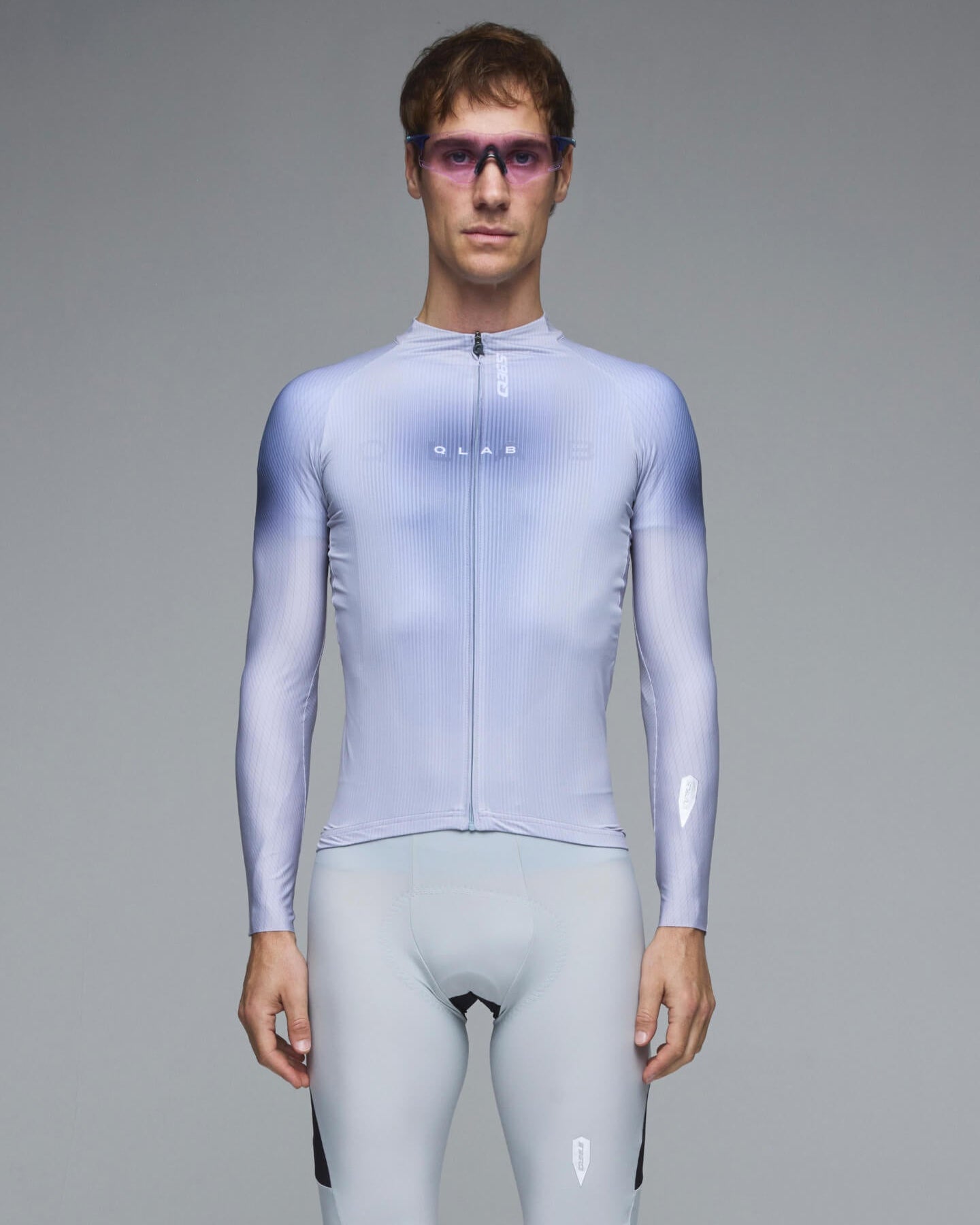 A male model wearing a light blue and white gradient long-sleeve cycling jersey and matching light blue cycling shorts, standing against a solid gray studio background.