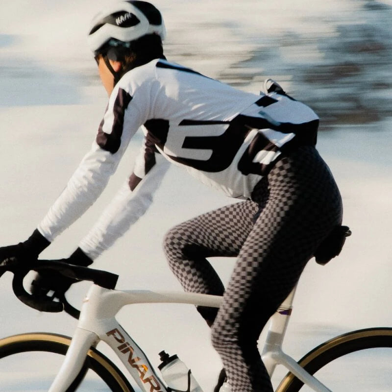 A cyclist in a white and black long-sleeve jersey, black-and-white patterned tights, and a white helmet rides a white Pinarello road bike on a snowy, blurred outdoor background.